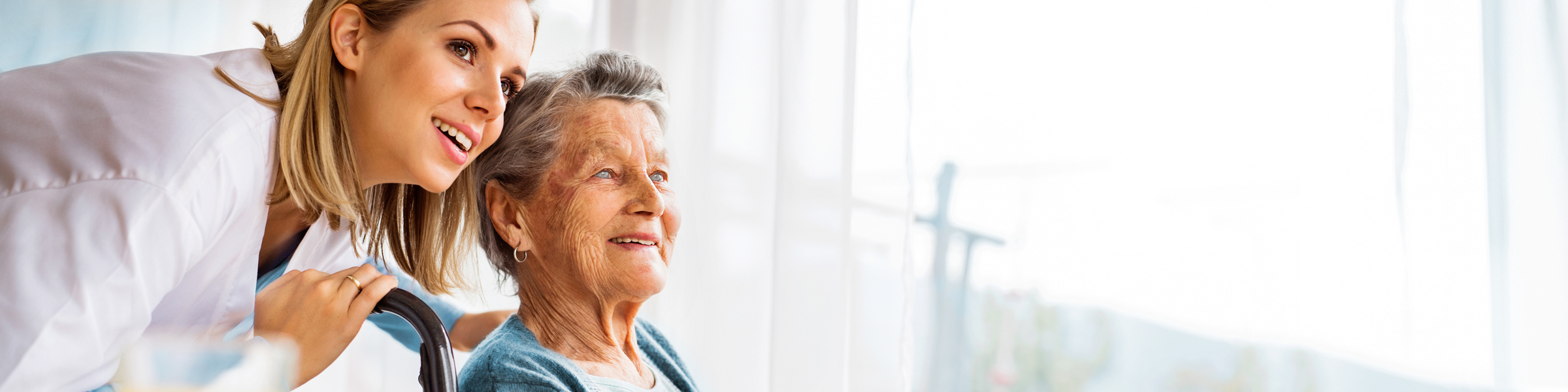 Startbild mit junger, lächelnder Betreuerin und einer blinden Bewohnerin, die beide aus dem Fenster blicken.